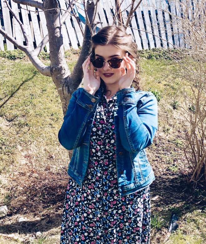 Julianne peers over the rims of her Ray Ban sunglasses. She is wearing a navy 90s midi dress with a floral pattern and a denim jean jacket.