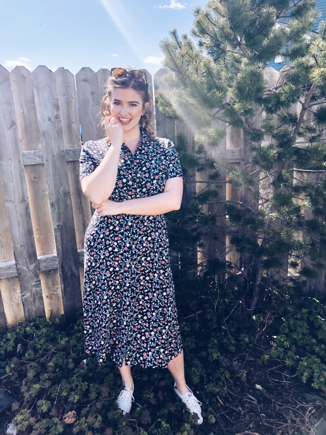 Julianne stands outside in front of a fence. She is wearing Ray Ban sunglasses on her head, white leather Keds shoes, and a navy 90s midi button up midi-dress with a floral pattern.